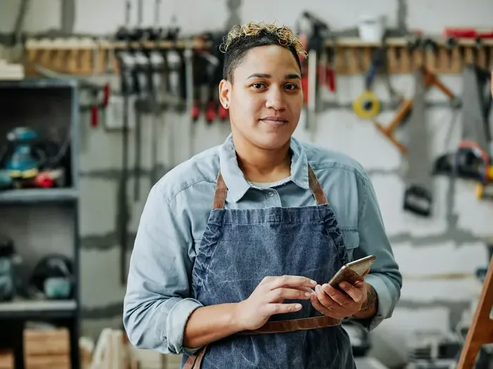 Local craftsman in apron standing in workshop holding phone surrounded by tools and wooden materials for Town Service Portal